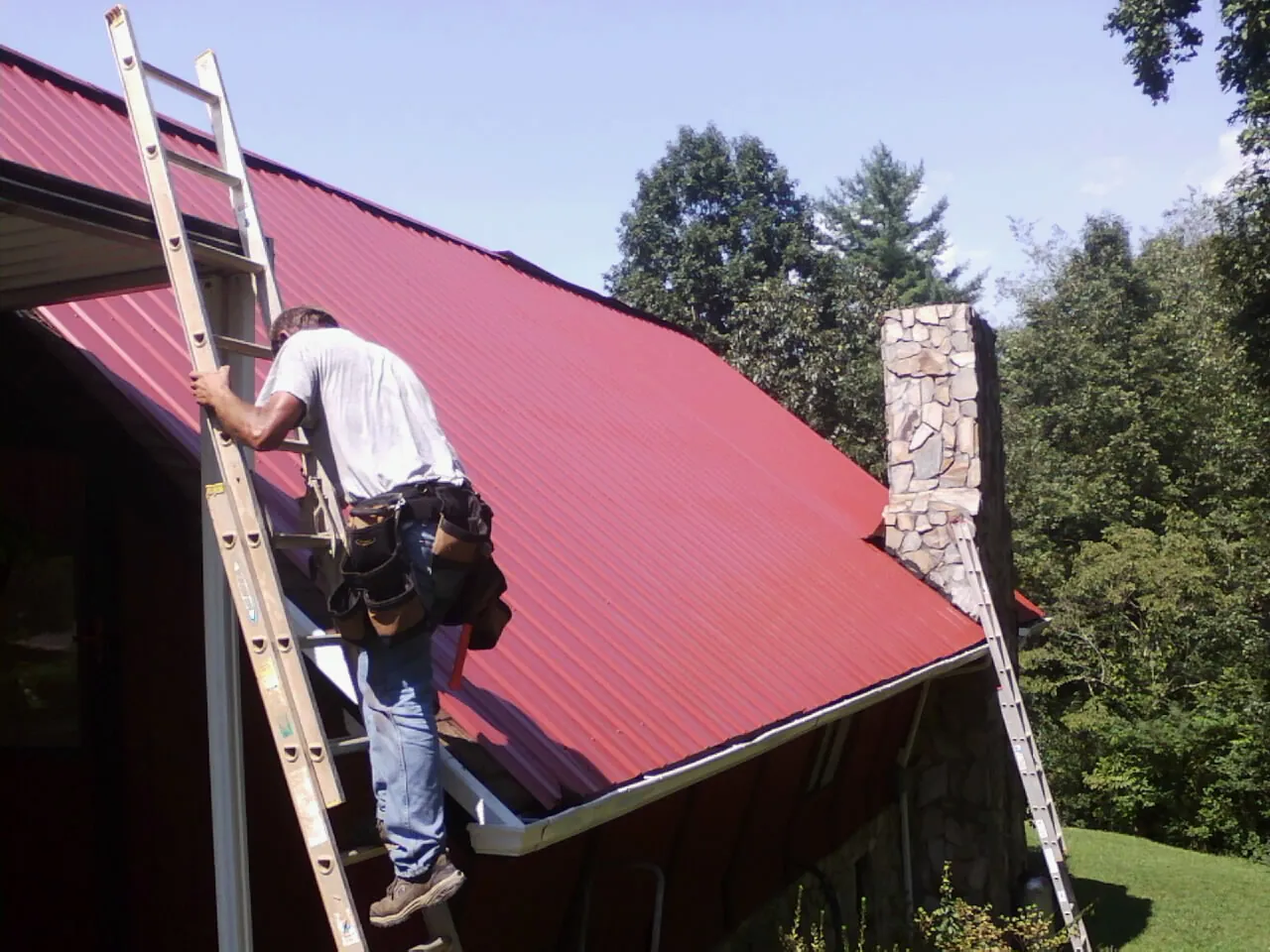 Roofer on a ladder beside a red metal roof and stone chimney — Soffit & Fascia Repair by Roofing Brothers in Richards